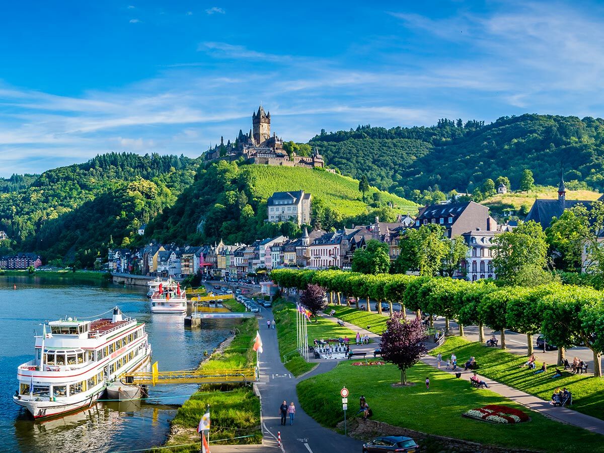 Panoramablick auf Cochem an der Mosel mit Reichsburg, Fluss, bunten Häusern und grünen Weinbergen unter blauem Himmel.