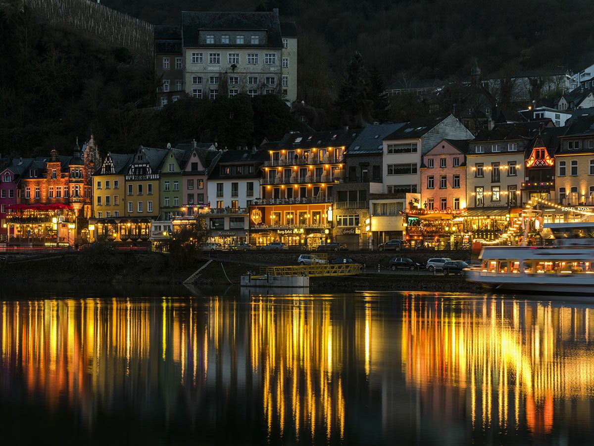 Malerische Moselstadt bei Nacht, mit bunten Häusern am Flussufer, Lichtern und Reflexionen im Wasser, und einem Schiff.