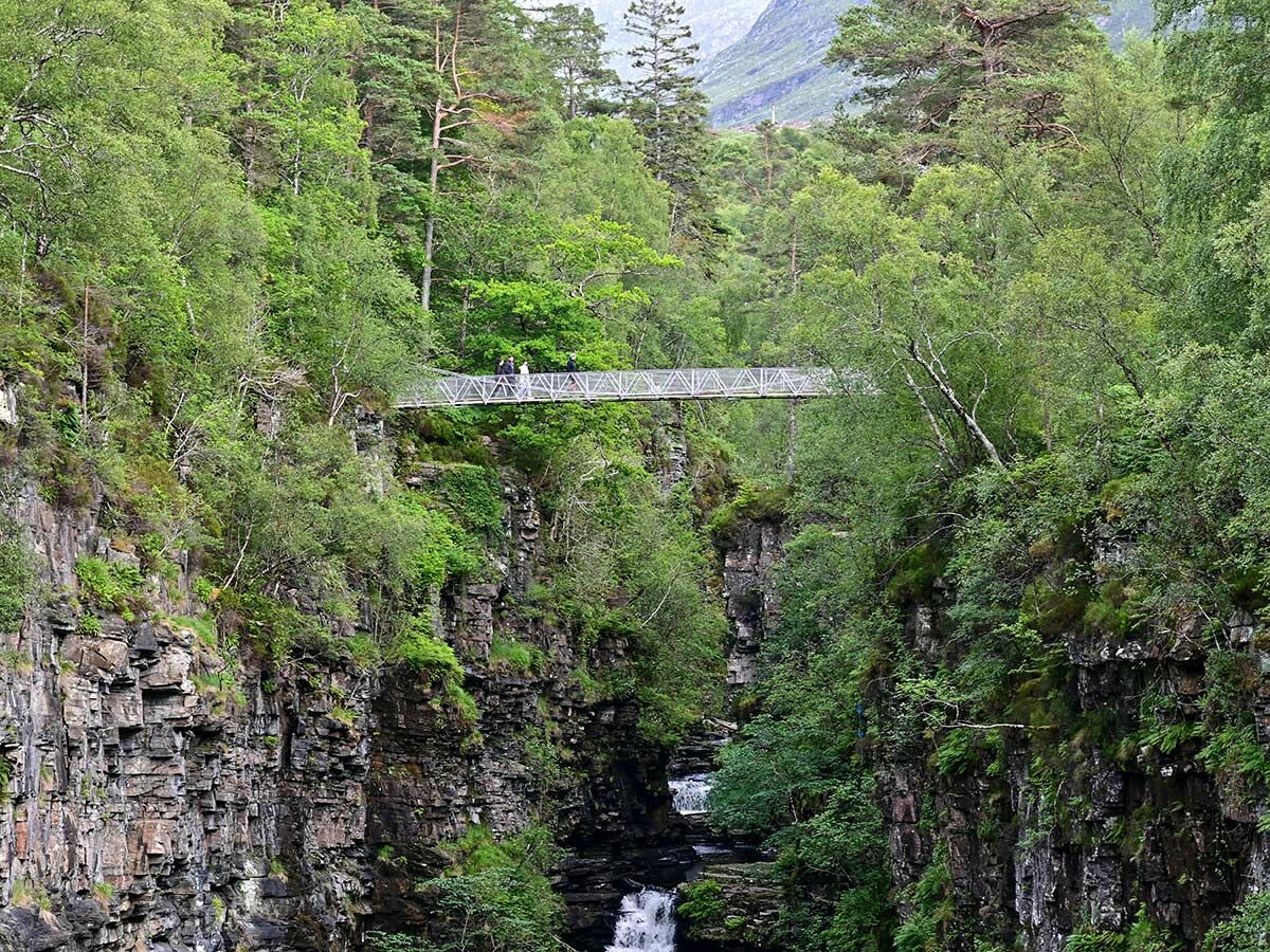 Hängebrücke überquert eine tiefe, bewaldete Schlucht mit Wasserfall, während Wanderer die malerische Natur genießen.