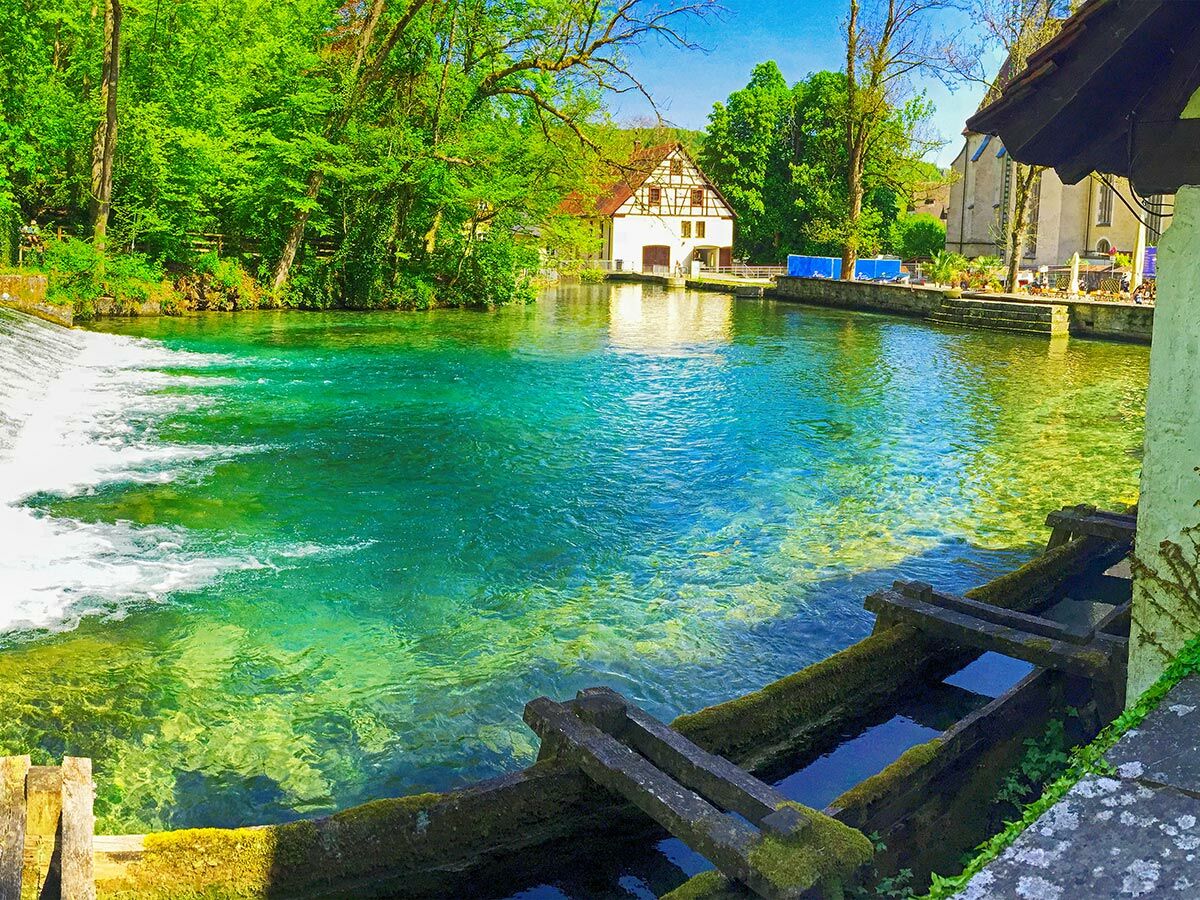 Idyllisches Dorf mit klarem, türkisgrünem Fluss, Wasserfall, grünen Bäumen und historischer Architektur am sonnigen Tag.