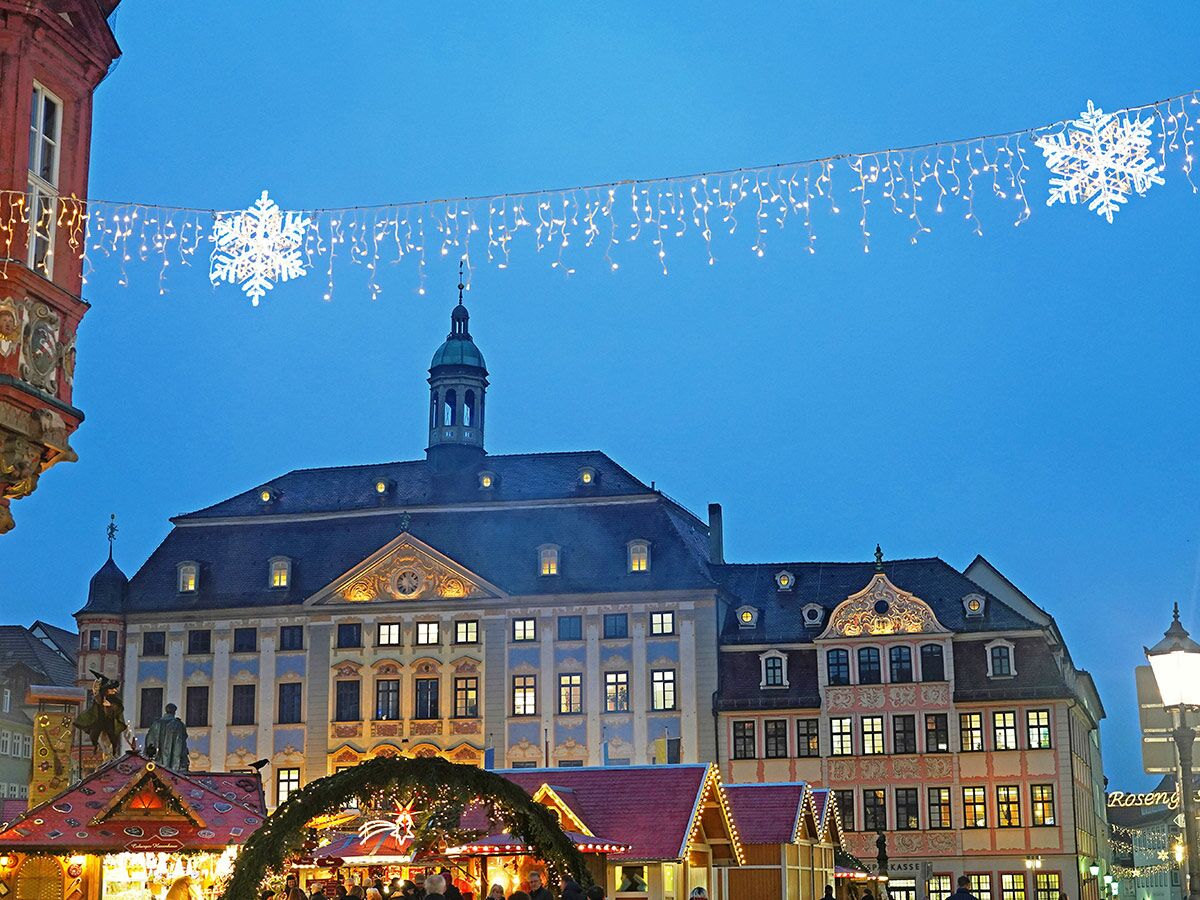 Weihnachtsmarkt bei Dämmerung in einer historischen deutschen Altstadt mit beleuchteten Gebäuden, Lichterketten und großen Schneeflocken.