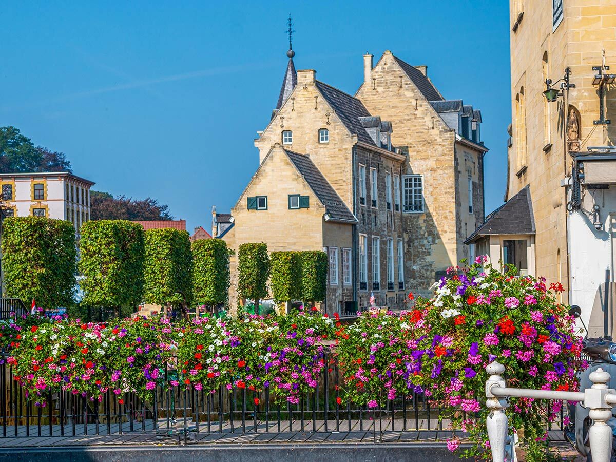 Malerische Ansicht von Maastricht mit historischen Gebäuden, blühenden Blumenkästen auf einer Brücke und blauem Himmel.