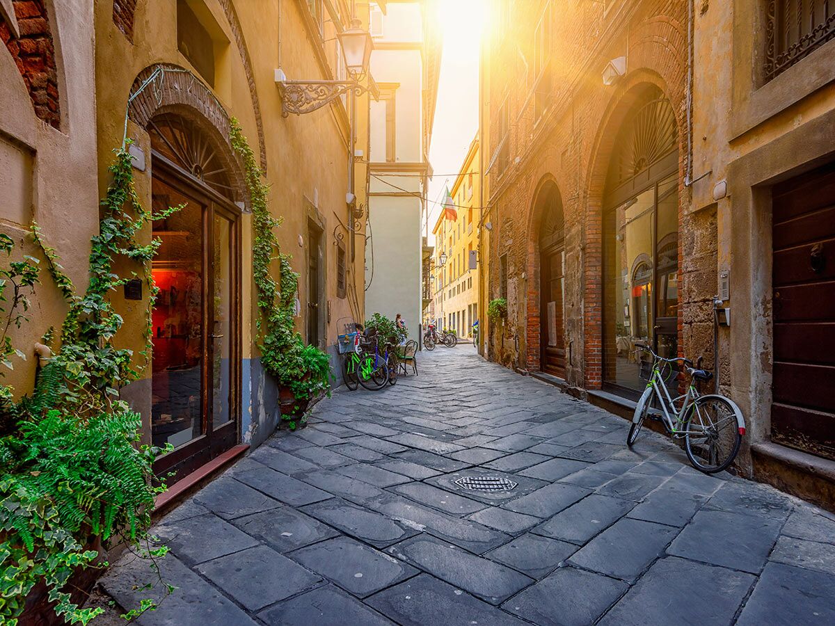Schmale Altstadtgasse in Italien mit historischen Gebäuden, geparkten Fahrrädern, Pflanzen und hellem Sonnenlicht am Ende.