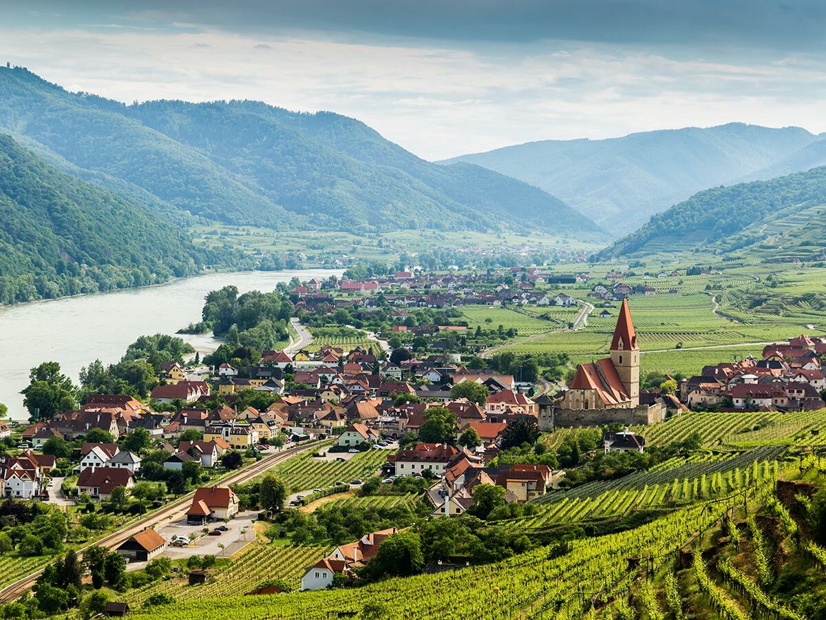 Luftbild von Weißenkirchen in der Wachau mit Donau, Weinbergen und der Pfarrkirche Maria Himmelfahrt unter blauem Himmel.