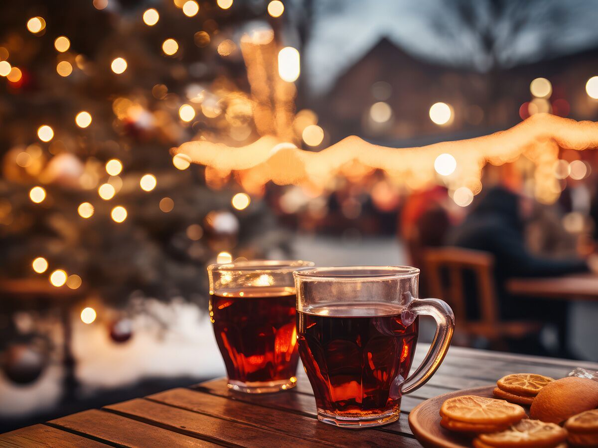 Zwei Glühweintassen auf Holztisch bei Weihnachtsbaum und Lichterketten auf einem winterlichen Weihnachtsmarkt im Abend.