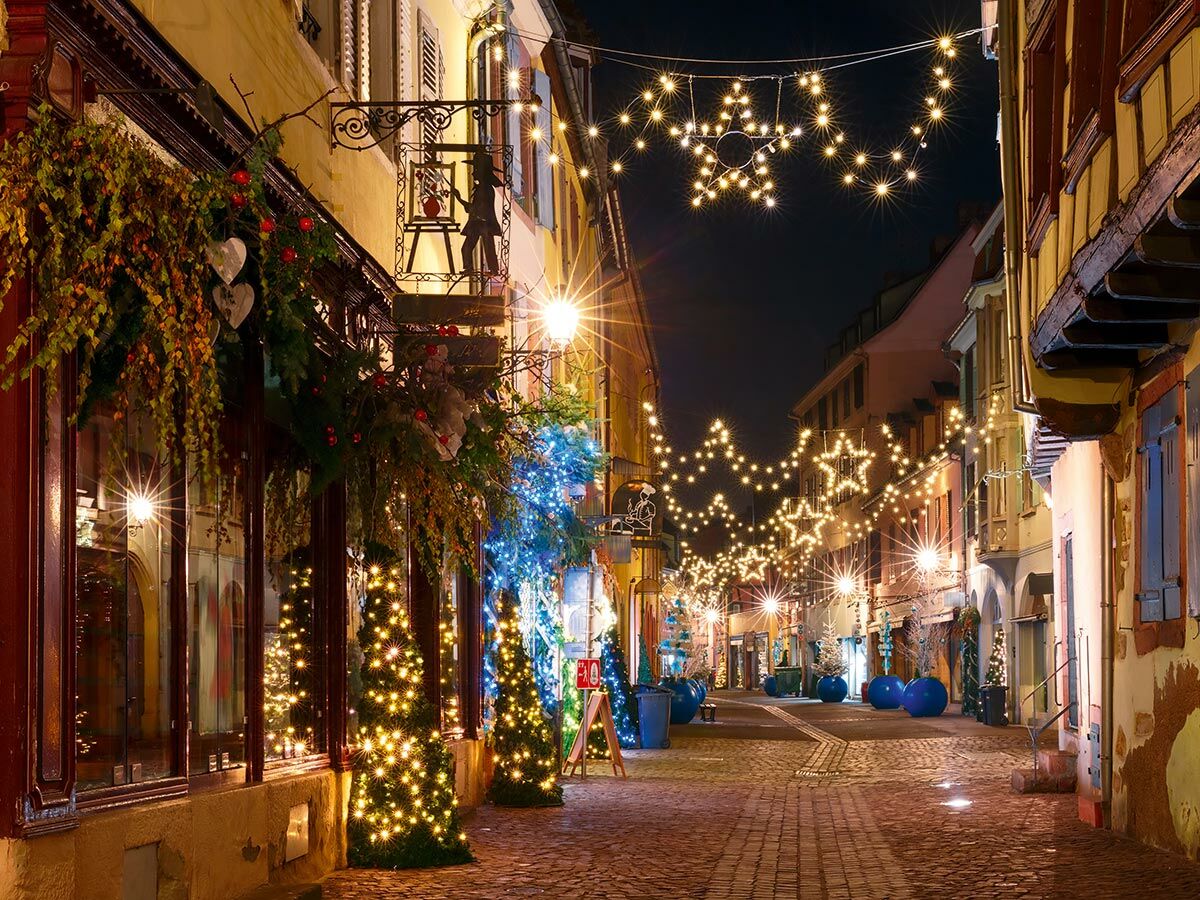 Festlich beleuchtete Straße in Colmar bei Nacht mit sternförmigen Lichterketten, Christbäumen und charmanten Geschäften.