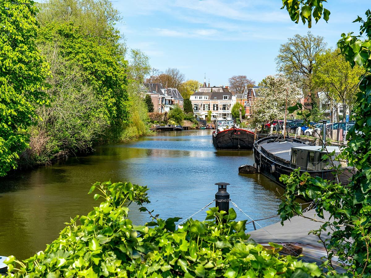 Ruhiger Kanal mit Hausbooten, gesäumt von grünen Bäumen und klassischen Häusern unter blauem Himmel.