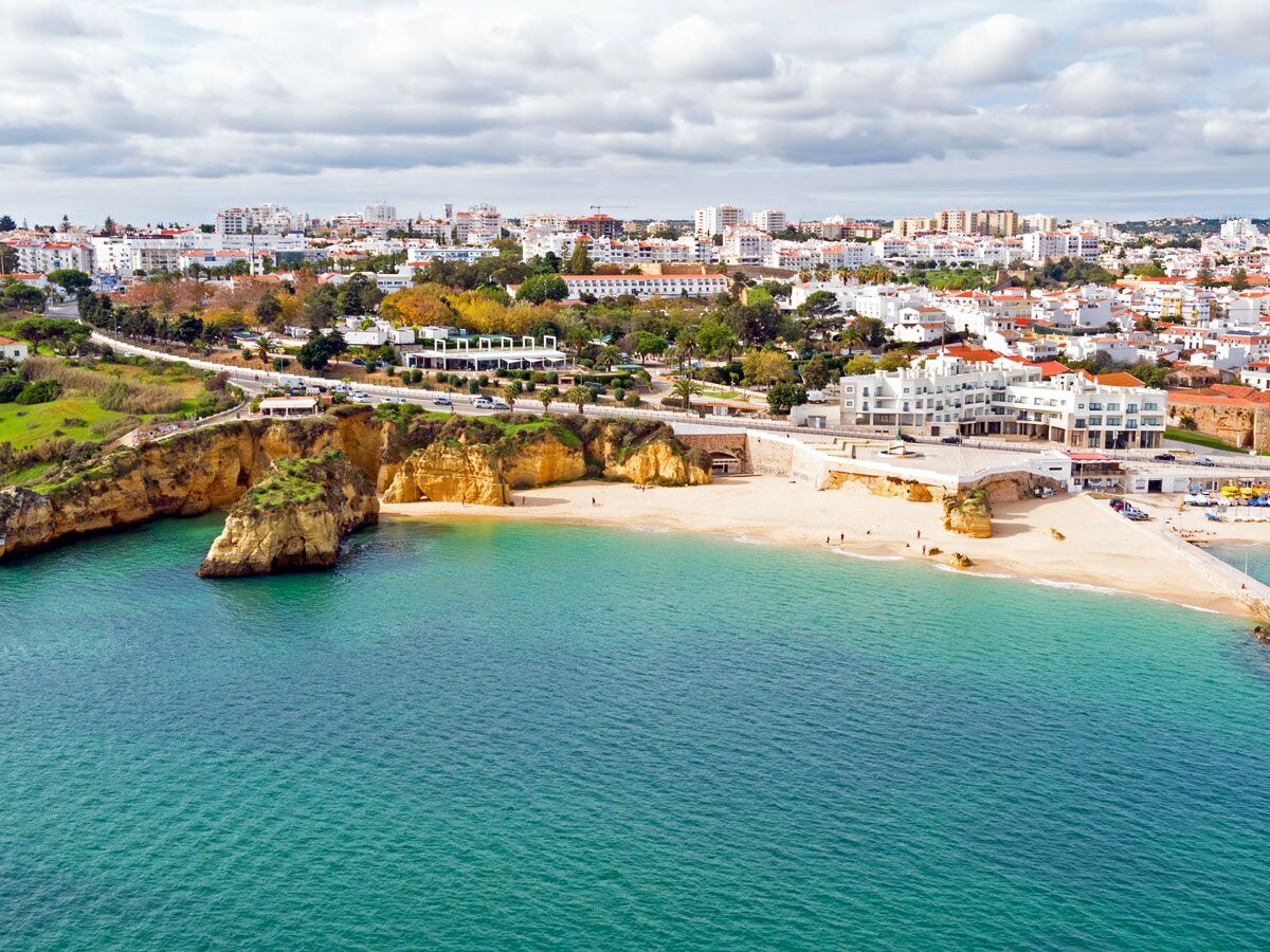 Luftbild einer malerischen portugiesischen Küstenstadt mit weißen Häusern, goldenen Felsen, sandigem Strand und türkisfarbenem Meer.