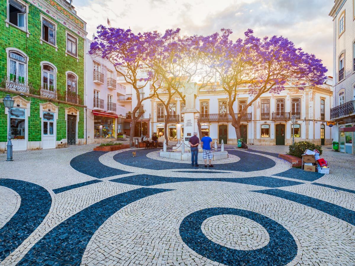 Historischer Marktplatz in Lagos, Portugal, mit lila blühenden Jacaranda Bäumen, traditioneller Architektur und Mosaikpflaster bei Sonnenuntergang.
