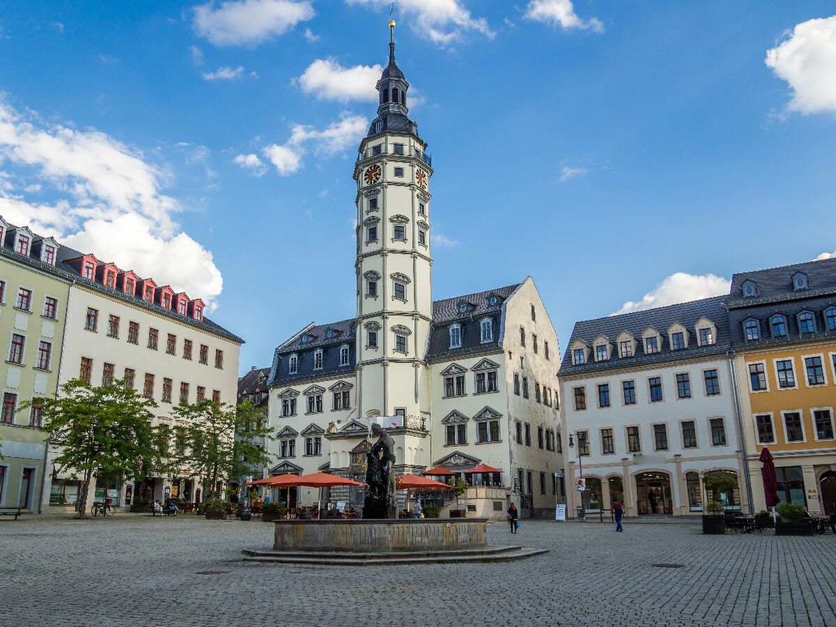 Historischer Marktplatz Oelsnitz/Erzgebirge mit Jakobikirchturm, Brunnen, Rathaus und Altstadthäusern unter blauem Himmel.