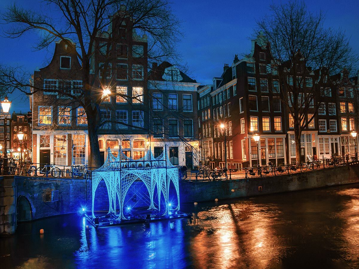 Nachtaufnahme der Amsterdamer Grachten mit beleuchteten historischen Häusern und blauer Lichtinstallation im Wasser.