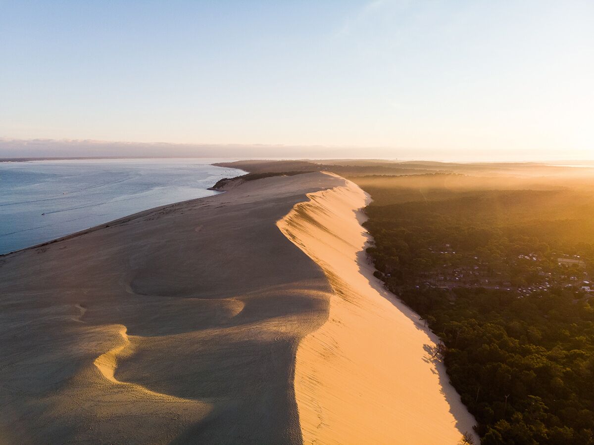 Luftaufnahme der riesigen Dune du Pilat bei Sonnenaufgang, mit Atlantik auf einer Seite und Kiefernwald auf der anderen.