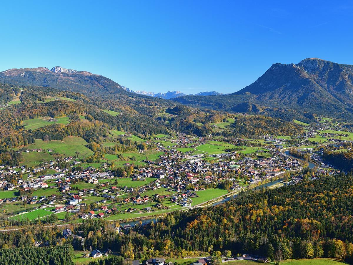 Weitwinkelaufnahme eines Alpendorfes im Herbst, umgeben von Bergen und Wäldern, mit einem Fluss durch das Tal.