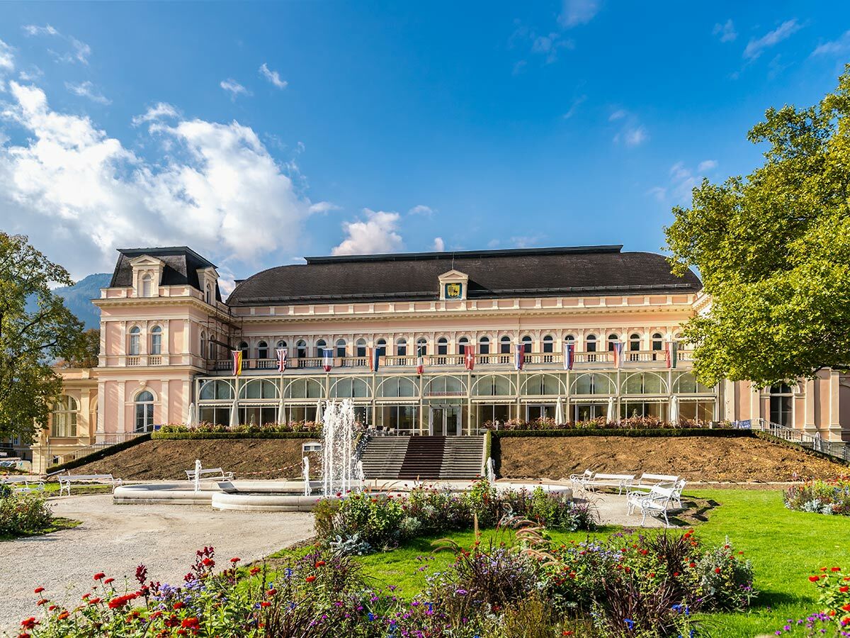 Elegantes Kurhaus mit rosafarbener Fassade, Garten, Springbrunnen, Blumen und Bergen im Hintergrund. Blauer Himmel.