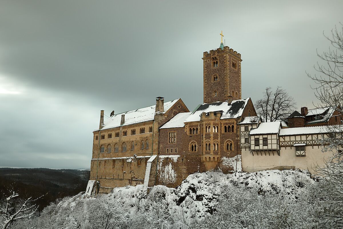 Majestätisches Wartburg Schloss im Winter, bedeckt mit Schnee, auf einem Hügel mit Blick auf verschneite Bäume unter bewölktem Himmel.