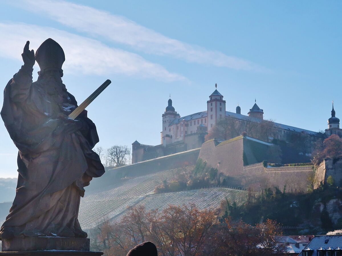 Bischofsstatue auf der Alten Mainbrücke mit Blick auf die winterliche Festung Marienberg in Würzburg unter blauem Himmel.