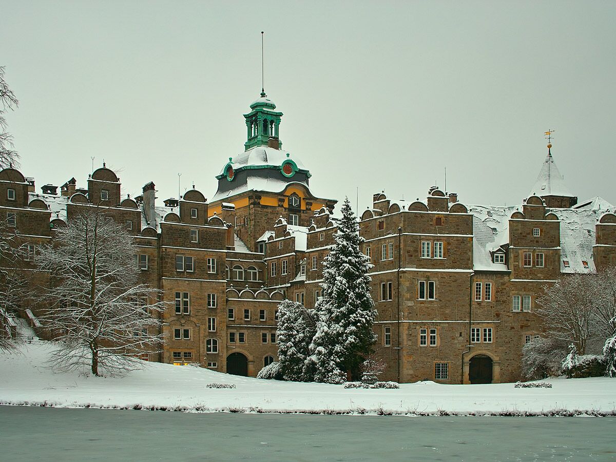 Imposantes steinernes Schloss mit schneebedeckten Dächern, einem grünen Kuppelturm und verschneiten Bäumen an einem gefrorenen See im Winter.