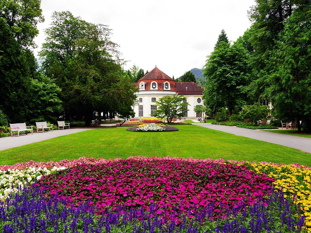 Prächtiger Kurgarten mit farbenfrohen Blumenbeeten, grünen Wiesen und einem eleganten, weißen Gebäude im Hintergrund.