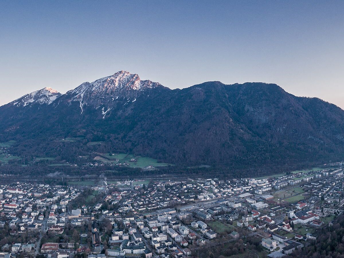 Luftaufnahme der österreichischen Stadt Salzburg mit Häusern und einem Fluss im Tal, vor imposanten, schneebedeckten Alpen.