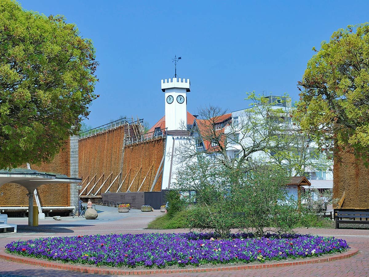 Gradierwerk mit Uhrenturm, leuchtendem Blumenbeet und grünen Bäumen in einem Kurpark an einem sonnigen Frühlingstag.