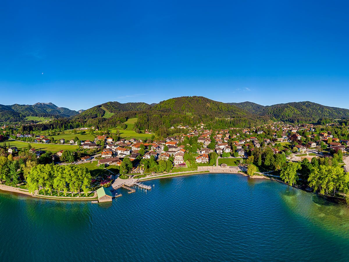 Luftbild eines idyllischen Dorfes am Tegernsee mit roten Dächern, grünen Bäumen und majestätischen Alpen im Hintergrund bei strahlendem Himmel.