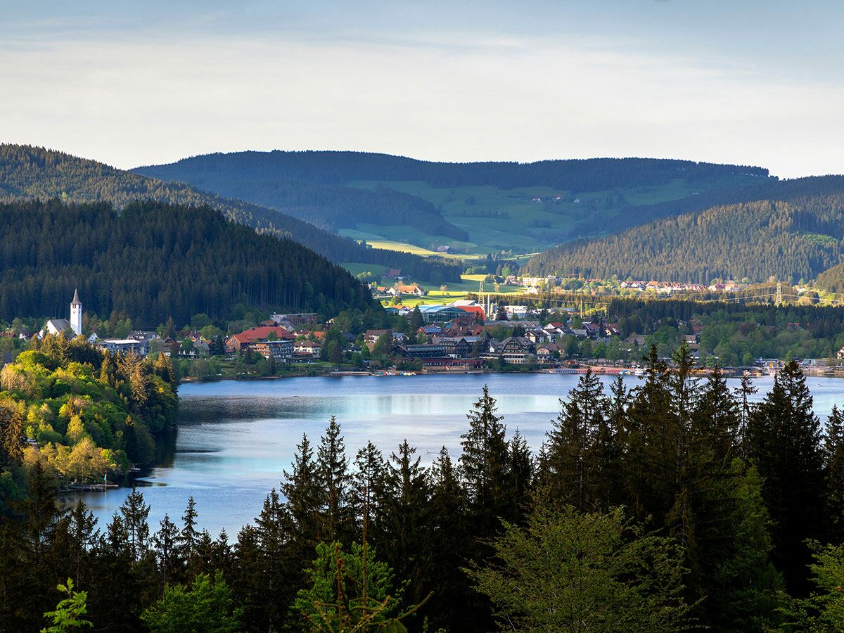 Panoramablick auf einen idyllischen See mit Dorf, weißer Kirche und bewaldeten Bergen im Schwarzwald, Deutschland.