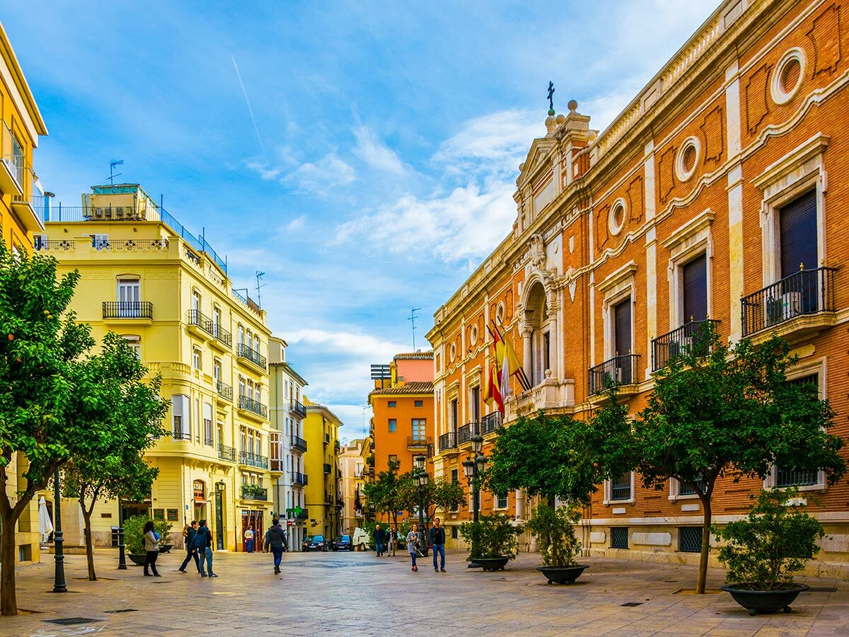 Historischer Stadtplatz in Spanien mit gelben und orangefarbenen Gebäuden, Menschen gehen unter blauem Himmel und Orangenbäumen.