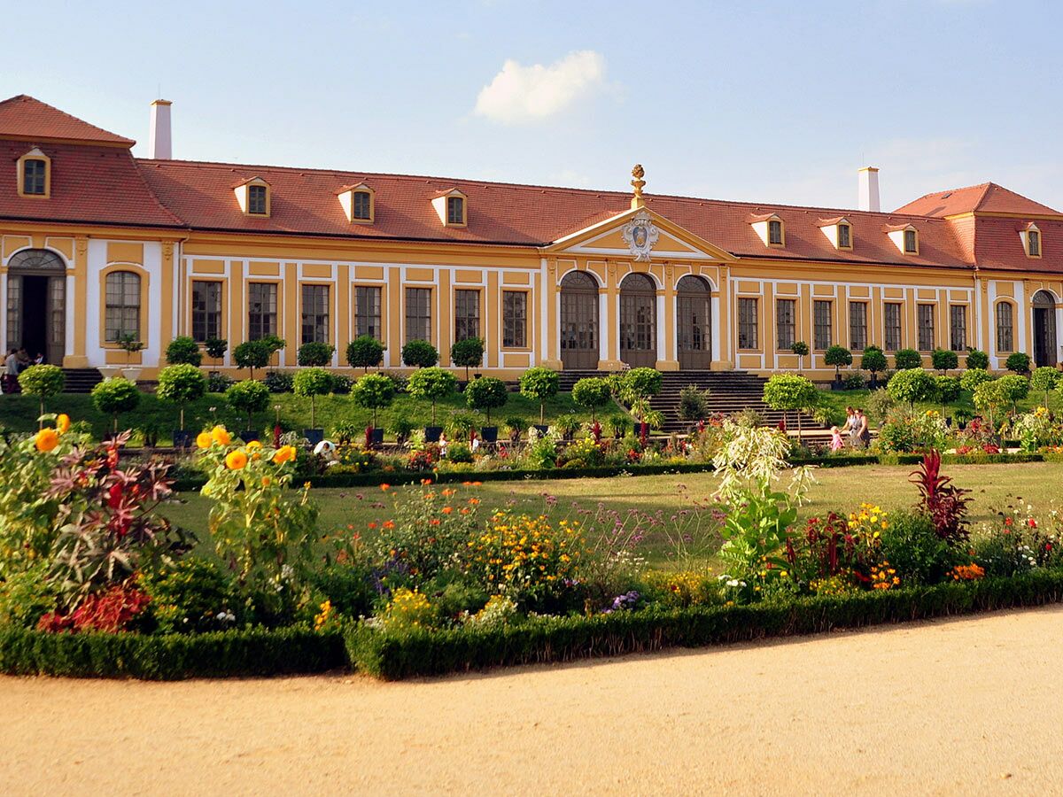 Gelbes Barockschloss mit rotem Ziegeldach. Davor ein weitläufiger, blühender Garten mit Zierbäumen unter blauem Himmel.