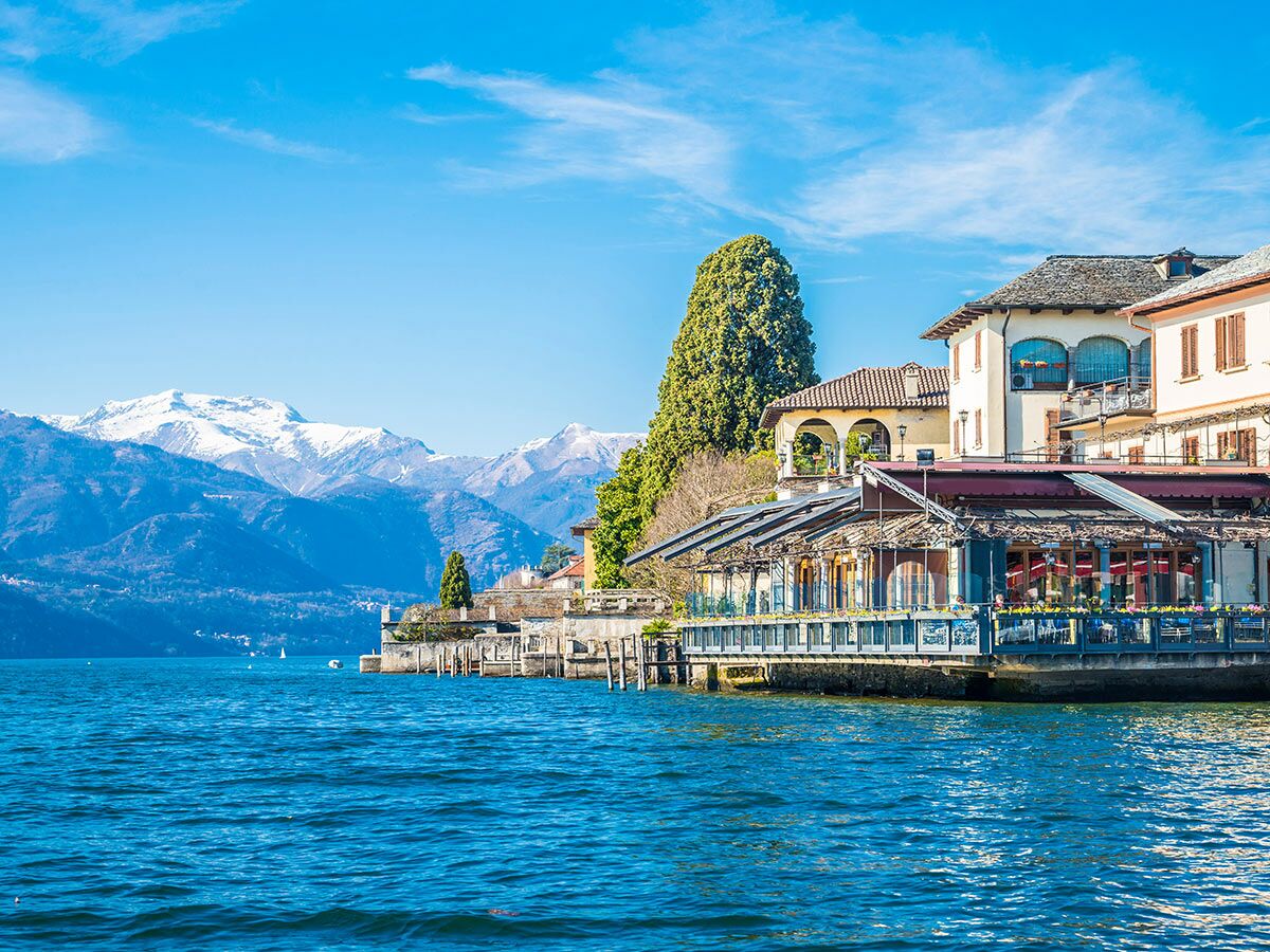 Malerischer Blick auf einen blauen See, schneebedeckte Alpenberge und charmante Gebäude mit einem Restaurant direkt am Ufer unter klarem Himmel.