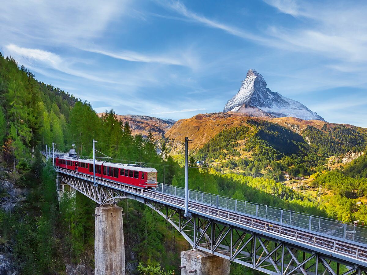 Roter Gornergratbahn Zug fährt über eine Brücke mit dem majestätischen Matterhorn und grünen Wäldern im Hintergrund.