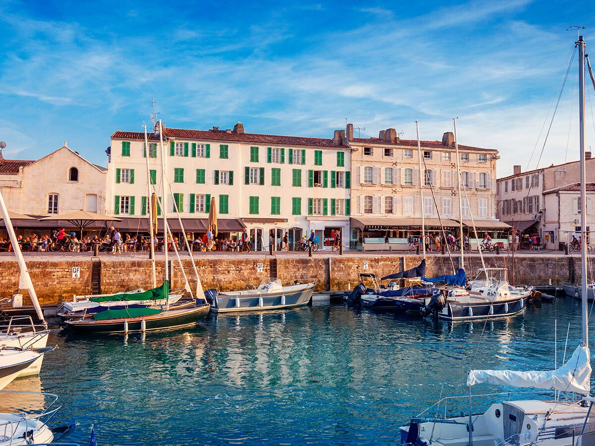 Malerische Hafenpromenade mit Cafés, historischen Gebäuden mit grünen Fensterläden und Yachten im Sonnenlicht.