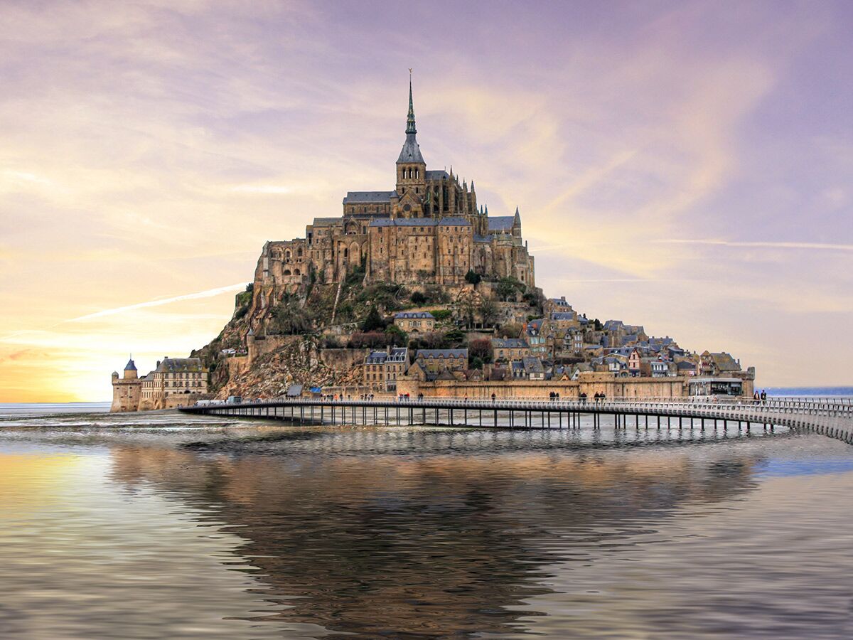 Mont Saint-Michel, eine beeindruckende Abtei auf einer felsigen Insel in Frankreich, mit einem Dorf und einer Brücke bei Sonnenuntergang und Spiegelung im Wasser.