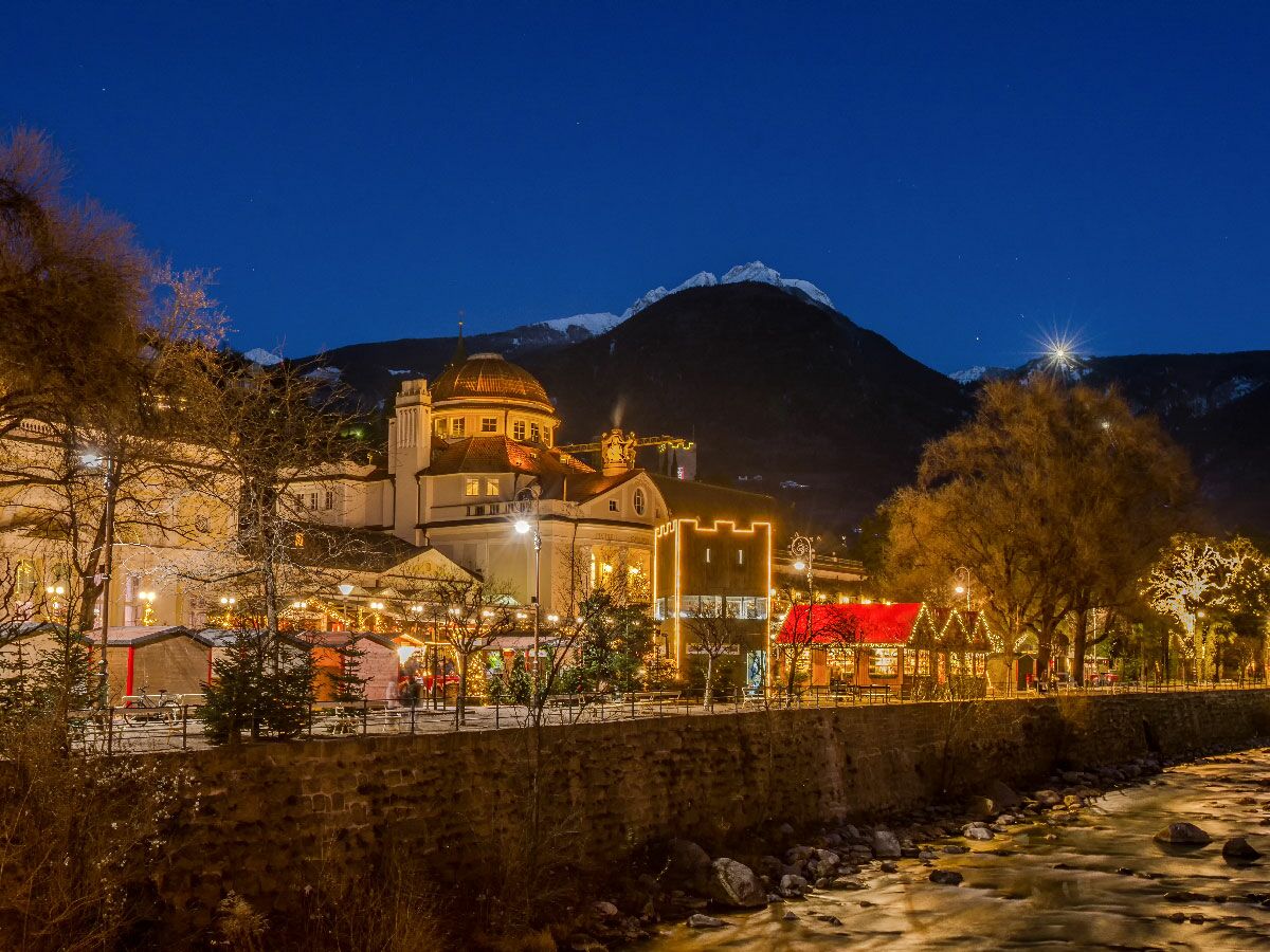 Winterlicher Weihnachtsmarkt in Meran am Fluss Passer bei Nacht mit beleuchteten Ständen, Kurhaus und verschneiten Bergen.