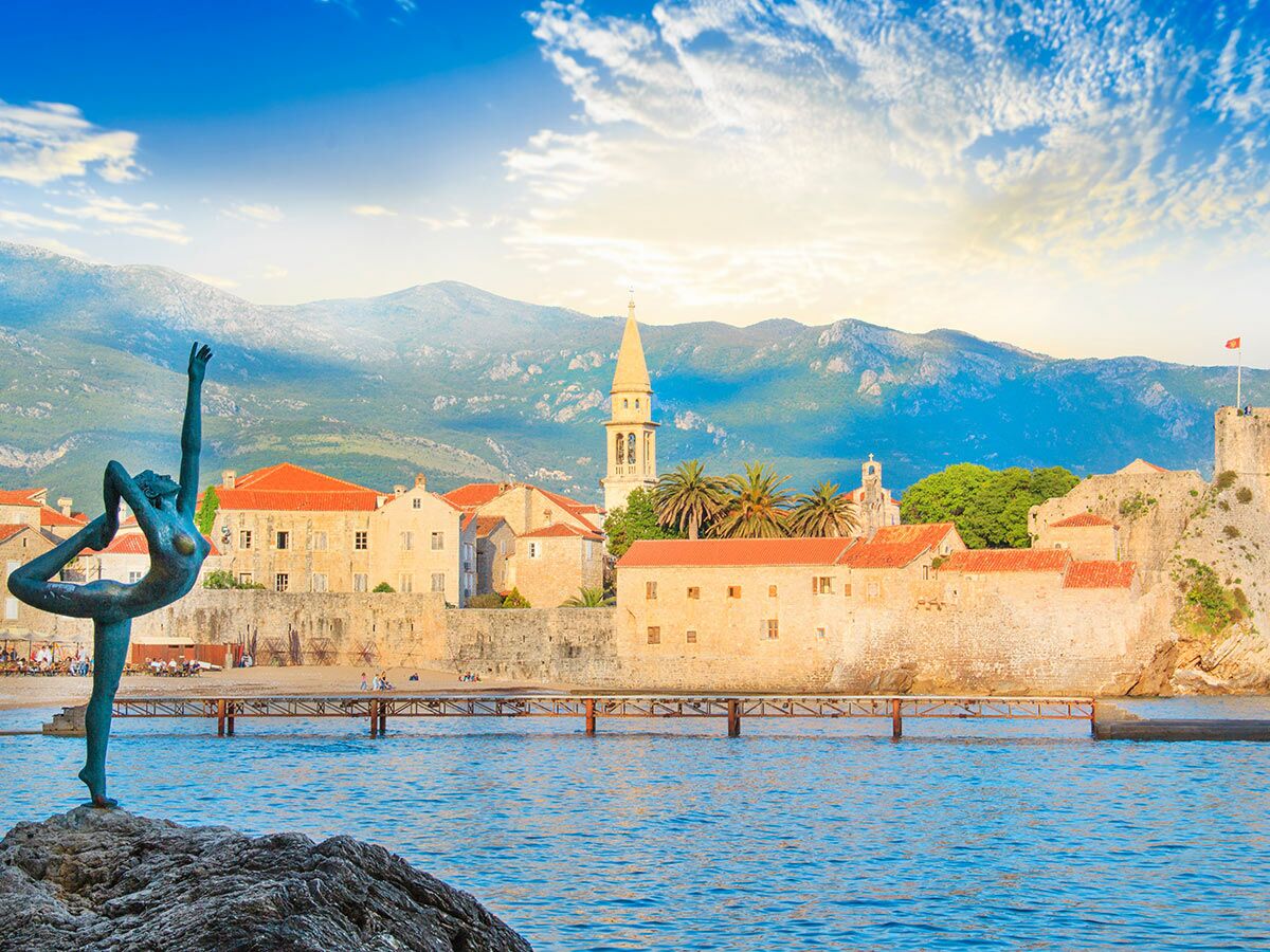 Bronzestatue Tänzerin auf Felsen mit historischer Altstadt von Budva, roten Dächern, Glockenturm, Meer und Bergen.