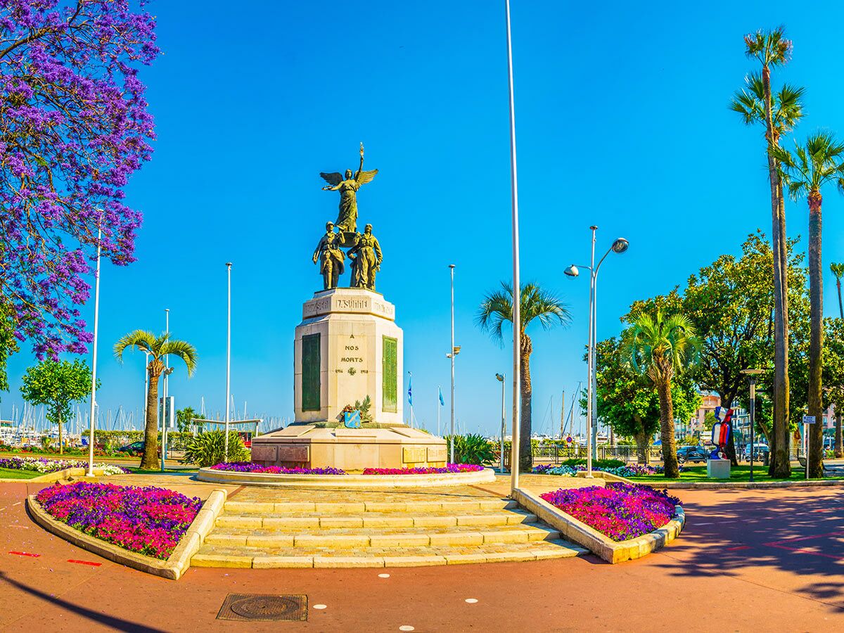 Kriegsdenkmal in Cannes mit geflügelter Statue und Soldatenfiguren, umgeben von blühenden Jacaranda-Bäumen und Palmen am Yachthafen.