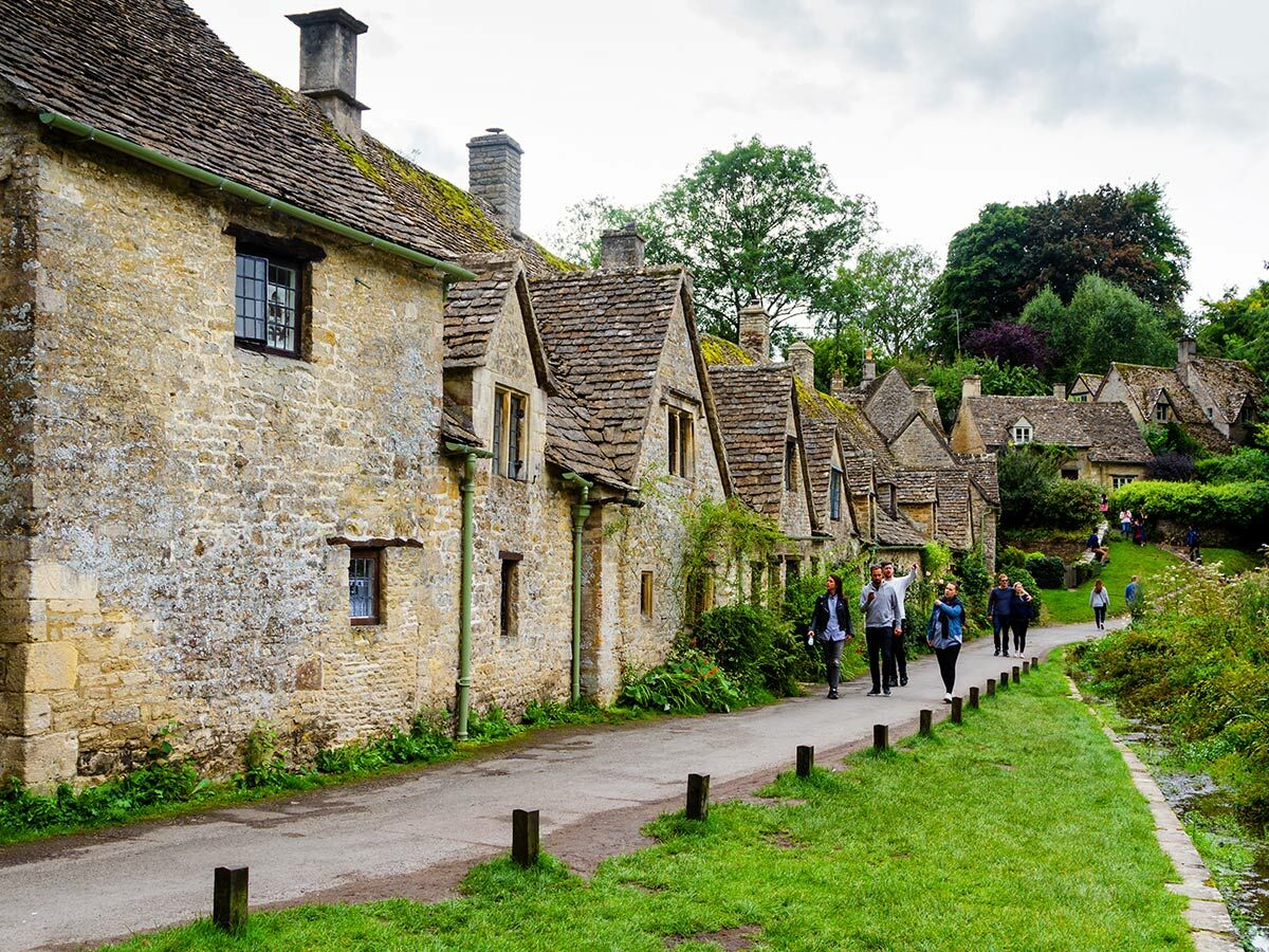 Idyllisches Bibury Dorf in den Cotswolds mit historischen Steinhäusern, moosbewachsenen Dächern und Spaziergängern am Weg.