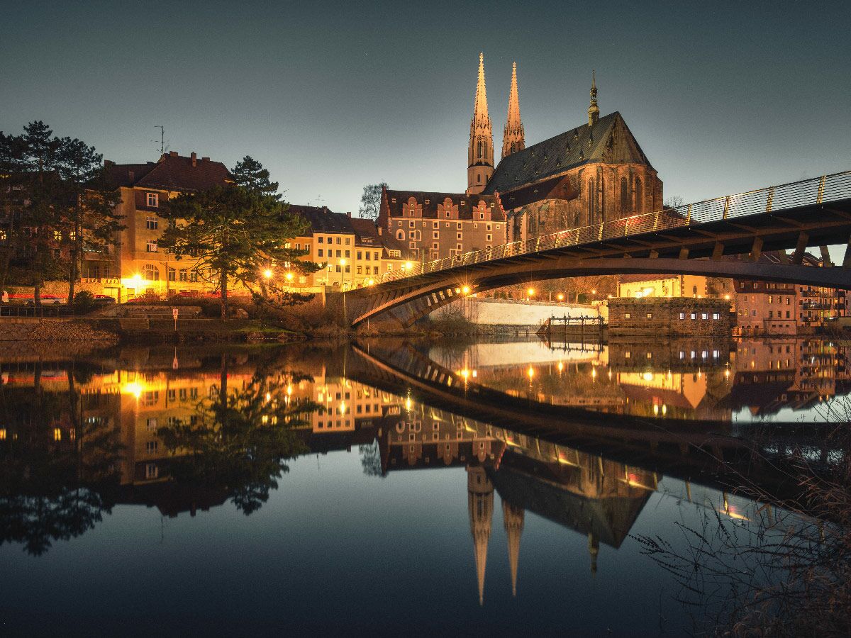 Nachtaufnahme der beleuchteten Peterskirche und Altstadtbrücke in Görlitz, mit Spiegelung im Fluss Neiße.