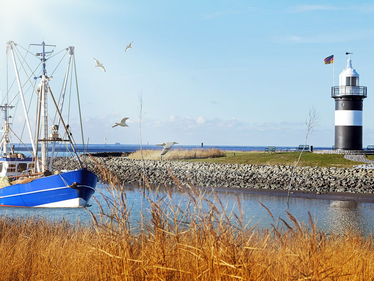 Blaues Fischerboot fährt vorbei an schwarz-weißem Leuchtturm und fliegenden Möwen an der deutschen Küste.