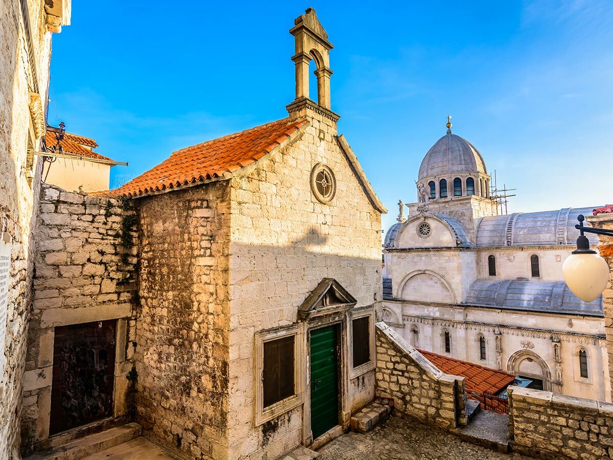 Historische Kathedrale des Heiligen Jakob in Šibenik, Kroatien, mit kleiner Steinkirche und Altstadt unter blauem Himmel.