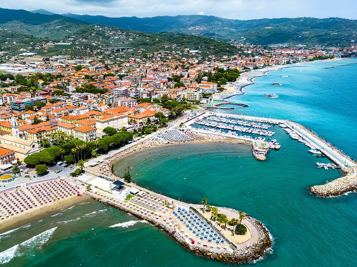 Luftaufnahme einer lebhaften italienischen Küstenstadt mit Yachthafen, Stränden, Gebäuden mit roten Dächern und grünen Hügeln.