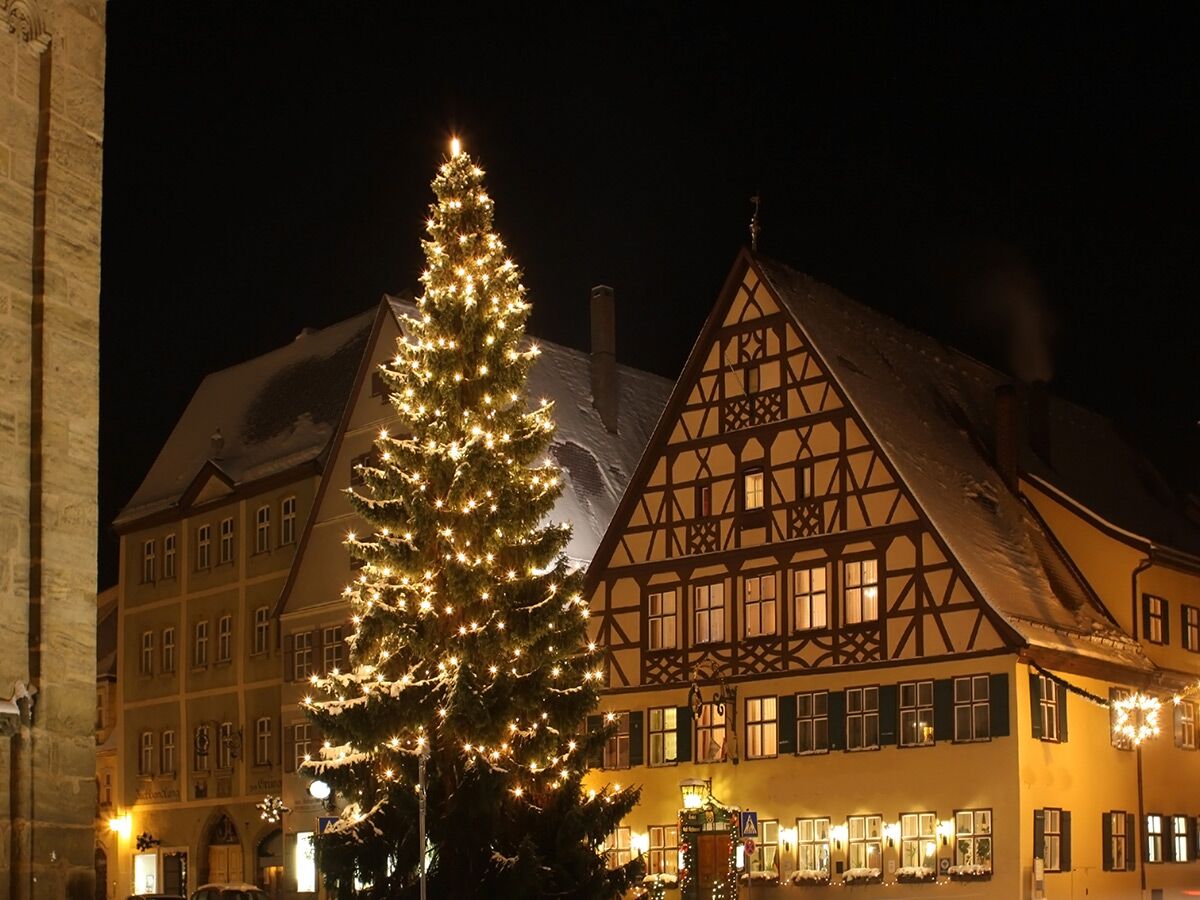 Großer Weihnachtsbaum leuchtet vor schneebedeckten Fachwerkhäusern in einer historischen deutschen Altstadt bei Nacht.
