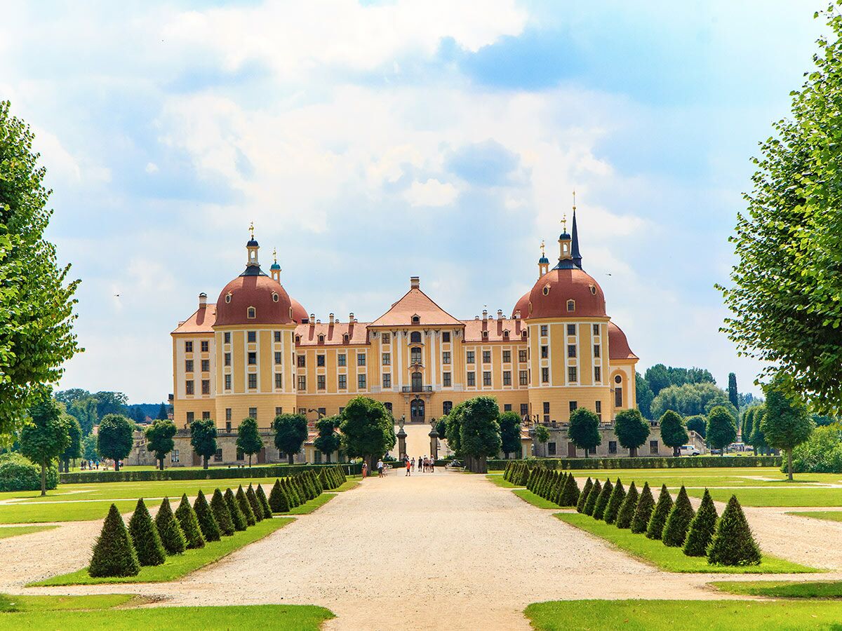 Blick auf das gelbe Barockschloss Moritzburg mit roten Dächern, umgeben von einem großen Park mit Hecken und Wegen bei Sonnenschein.