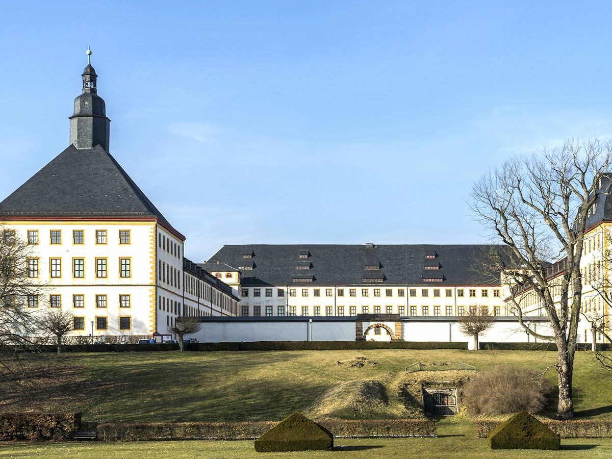 Panoramablick auf ein imposantes Barockschloss mit hohem Turm, umgeben von einem grünen Park unter blauem Himmel.