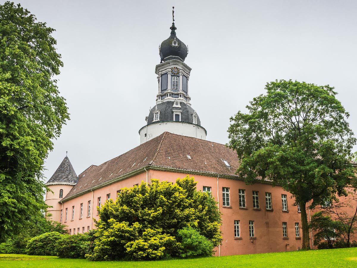 Historisches Schloss mit markantem Kuppelturm und rosafarbenem Gebäude, umgeben von grünen Bäumen und Rasen.
