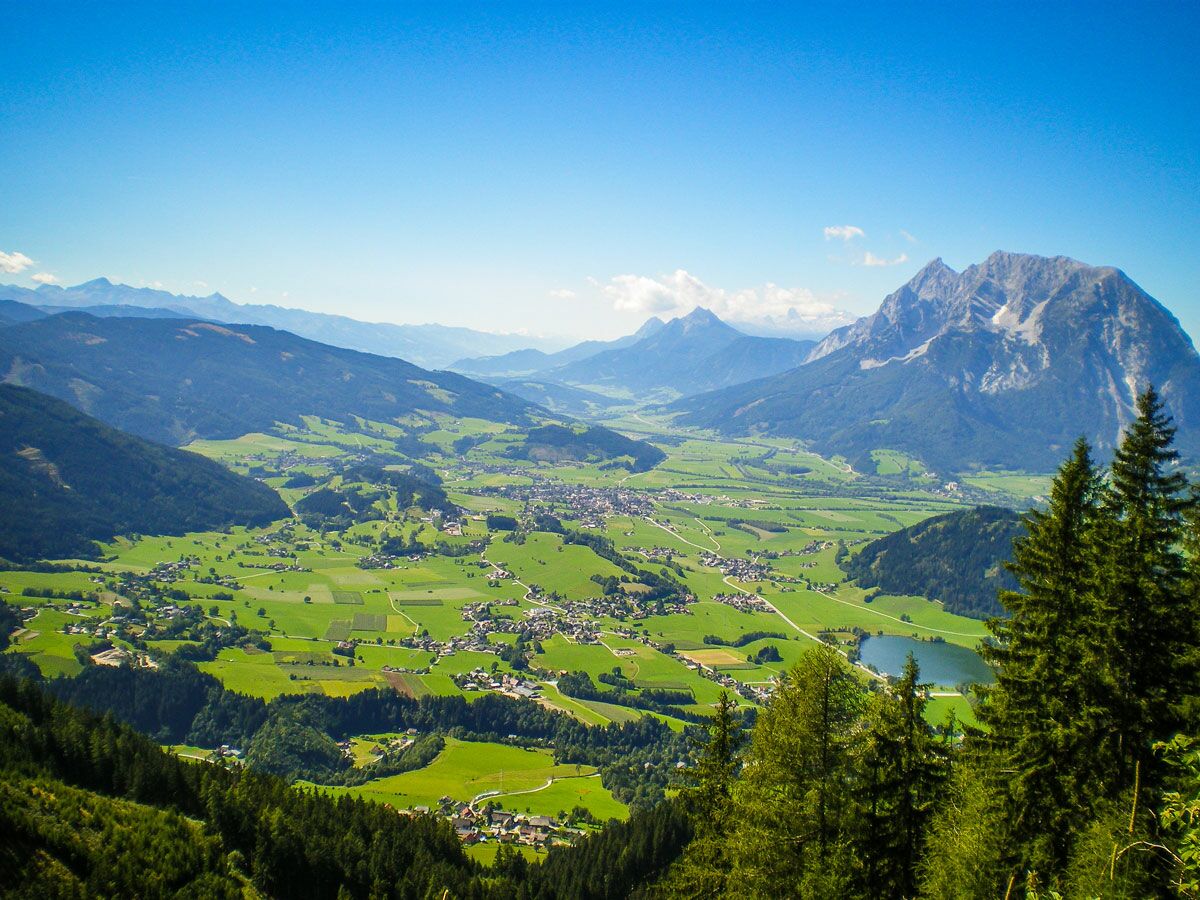 Panoramablick auf ein weites grünes Alpental mit Dorf, hohen Bergen, Wäldern und einem kleinen See unter blauem Himmel.