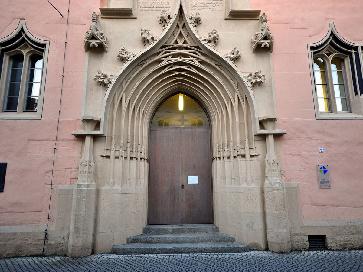 Prächtiger gotischer Eingang eines historischen Gebäudes mit Steinbogen, geschnitzten Details, Holztür, Bogenfenstern und rosa Fassade. Schild: Landeskirchenamt.