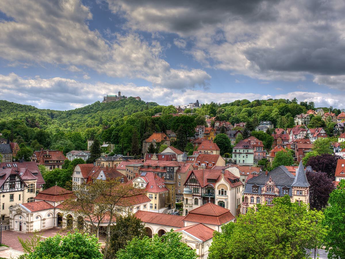 Panoramablick auf die historische Stadt Eisenach mit roten Dächern und grünen Bäumen. Das Schloss Wartburg thront auf einem bewaldeten Hügel.