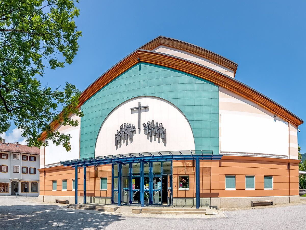 Moderne Kirche mit geschwungener Fassade in Türkis und Orange, großem Kreuz und stilisierten Figuren. Blauer Himmel, grüner Baum.