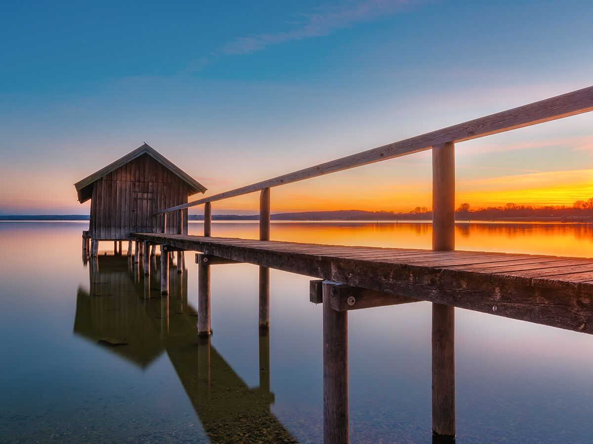 Idyllisches Holz-Seehaus mit Steg im See bei goldenem Sonnenuntergang mit perfekten Reflexionen.