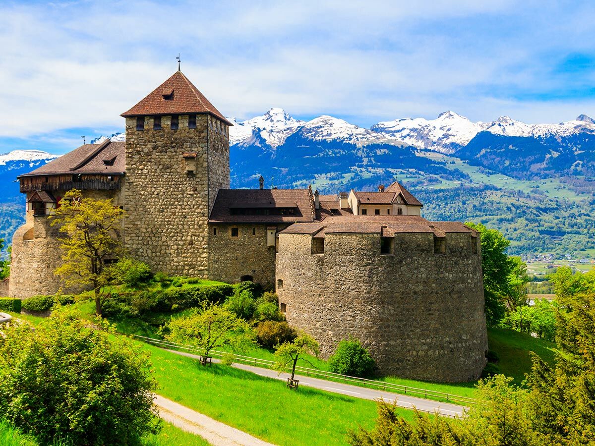 Historisches Schloss Vaduz mit steinerner Festung, Turm, grünen Hügeln und schneebedeckten Alpen unter blauem Himmel in Liechtenstein.